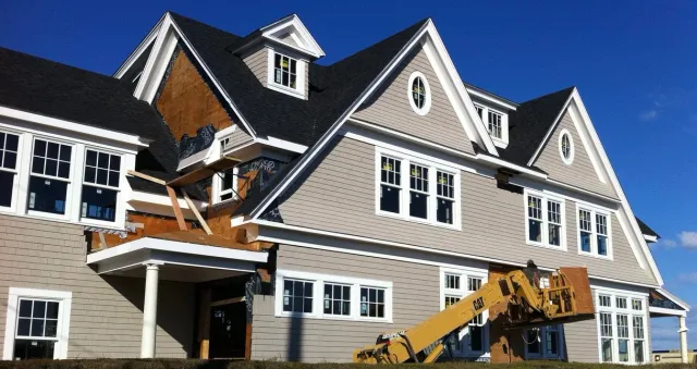 A large, multi-story house under construction with scaffolding and a lift. The house has beige siding, white window frames, and a black roof. Part of the exterior wall is unfinished, showing wooden framework and insulation.