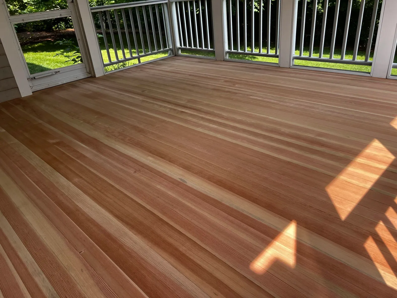 A newly installed wooden deck with light and dark wood planks, surrounded by white railing, with green grass and bushes visible outside.