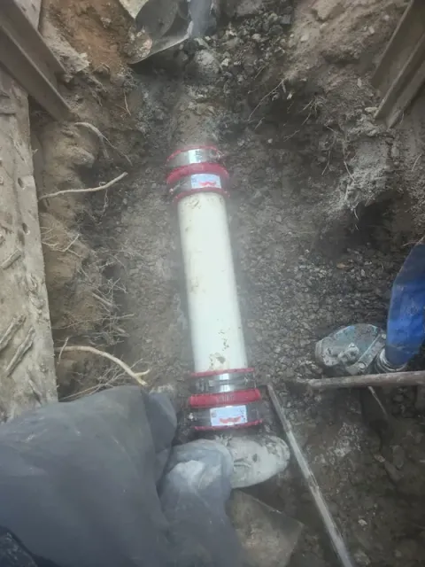Two workers installing a white pipe with red clamps in a muddy trench.