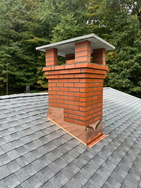 A red brick chimney with a concrete cap, rising from a grey shingled roof against a background of green trees.