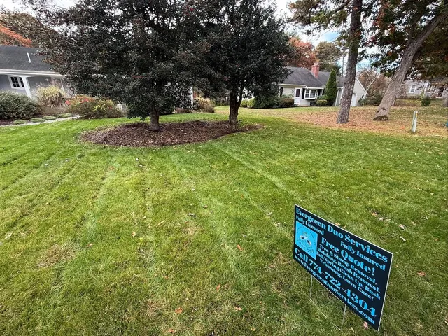 Green lawn with trees, houses in the background, a sign in the foreground with text and phone number.