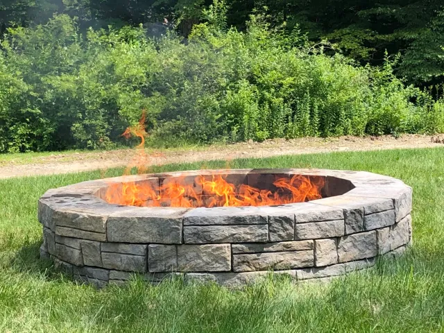 Stone fire pit with visible flames on green grass, background with bushes and trees.