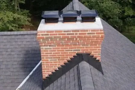 A brick chimney with three black metal caps sits on a dark shingled roof with stepped metal flashing.