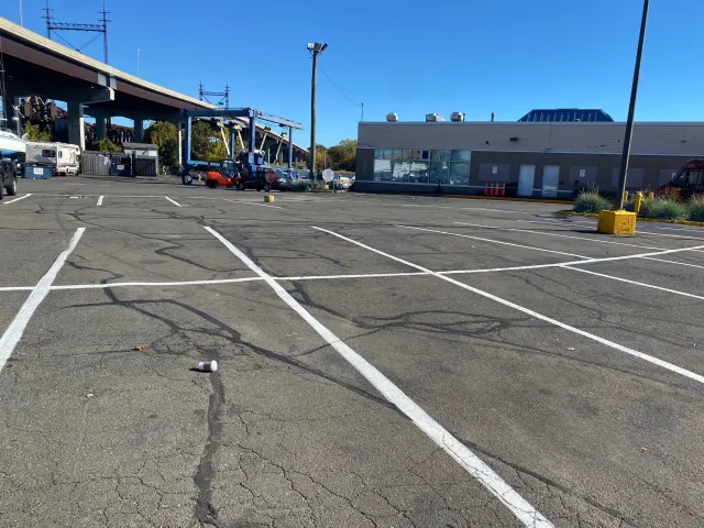 Empty parking lot with faint white lines, a discarded coffee cup, and a building with a large window and orange traffic cones in the background.