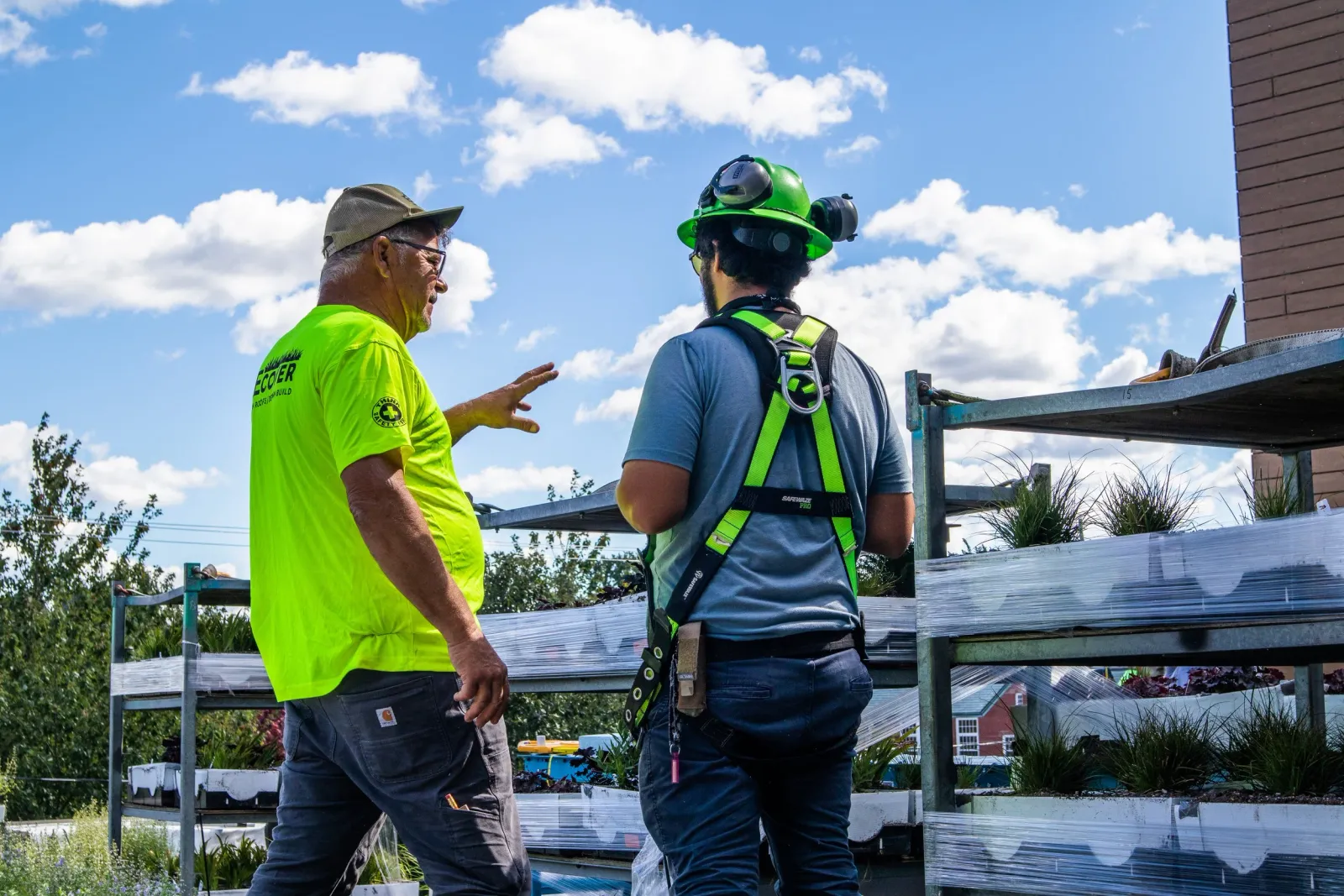 Two men are engaged in conversation on a rooftop garden with plants and flowers, both wearing safety gear and casual work clothing, under a blue sky with clouds.
