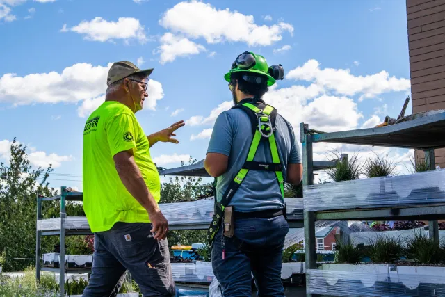 Two men are engaged in conversation on a rooftop garden with plants and flowers, both wearing safety gear and casual work clothing, under a blue sky with clouds.