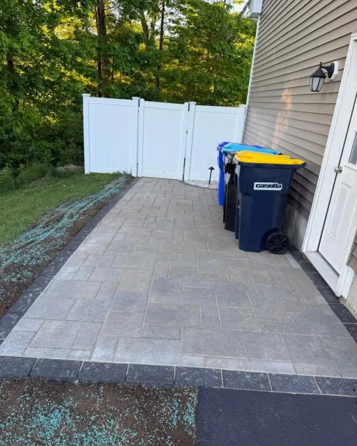 Paved backyard patio beside a house, with a white fence and blue trash bin.