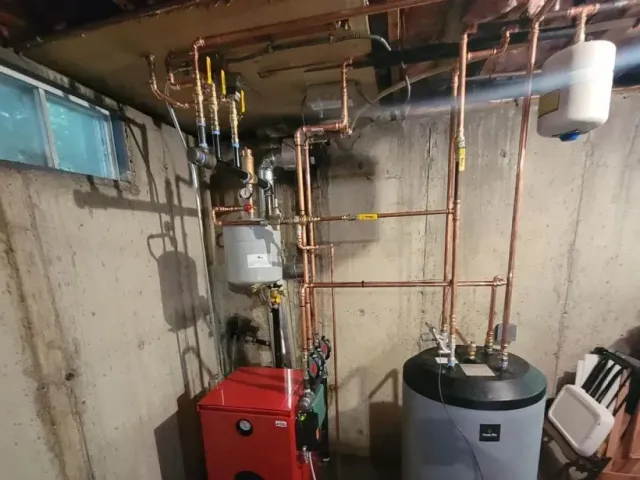 Basement mechanical room with red boiler, gray tank, and copper pipes along concrete walls