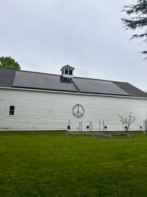 A white barn with solar panels on the roof, a small tree in front, a peace sign decoration on the wall, and a garden bed in the yard.
