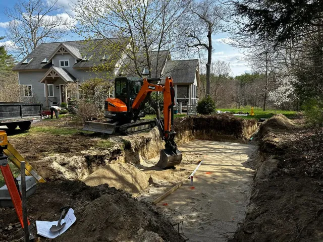 Excavator digging a rectangular foundation in a residential backyard with a house visible in the background.