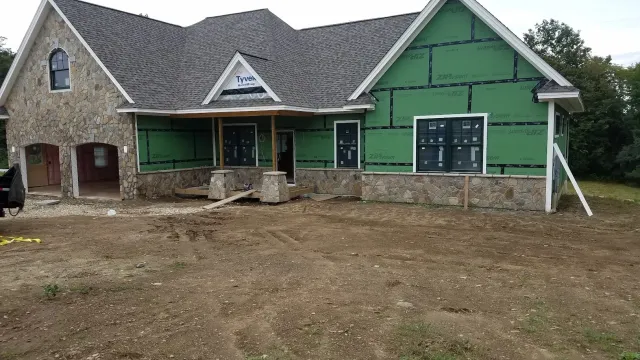 Under construction house with stone accents, green sheathing on part of the exterior, and a shingled roof. The front porch is unfinished, and the yard is dirt.