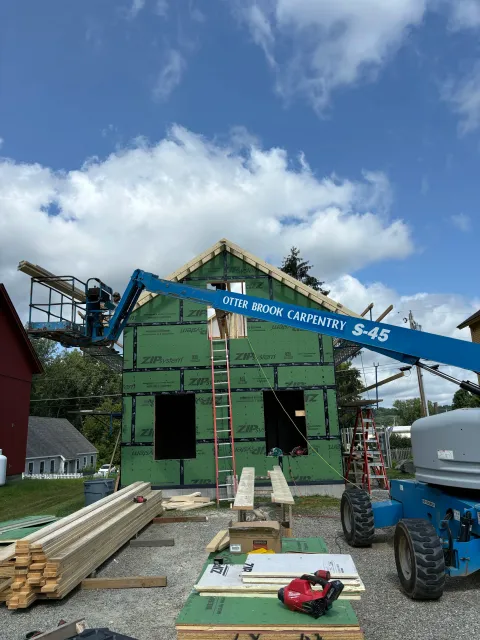 A house under construction with green sheathing and a blue man lift labeled 'Otter Brook Carpentry' lifting materials. Construction materials such as wood planks and tools are scattered around.