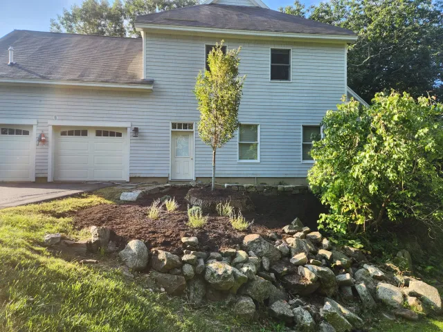 Front of a white house with a garage, a door, and multiple windows. A newly planted lean tree and shrubbery are visible in the yard. The yard has a rock border and some freshly disturbed soil.