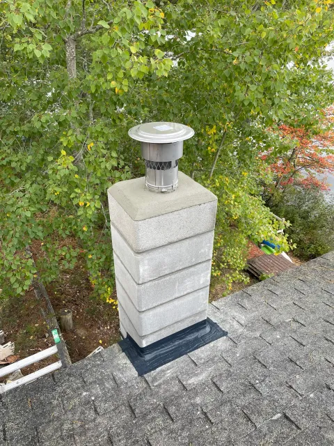 A metal chimney cap on top of a brick chimney on a shingled roof, with green leafy trees in the background.