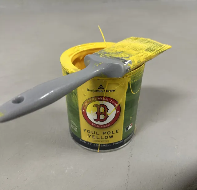 A partially opened tube of Benjamin Moore yellow paint with yellow paint smeared on the cap and the brush resting on top, set against a light gray background.