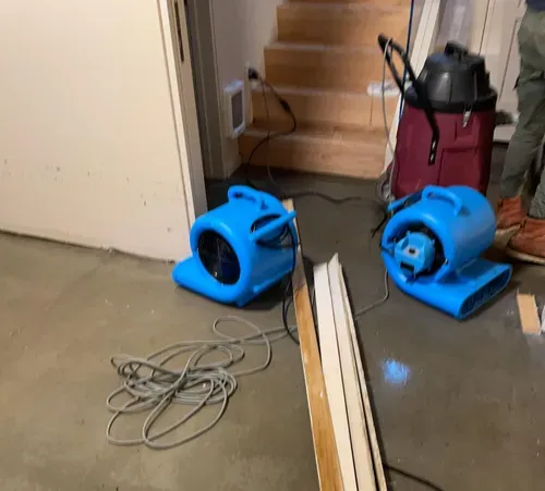 Two blue air movers and a red vacuum on a concrete floor near a staircase, with cords and boards nearby
