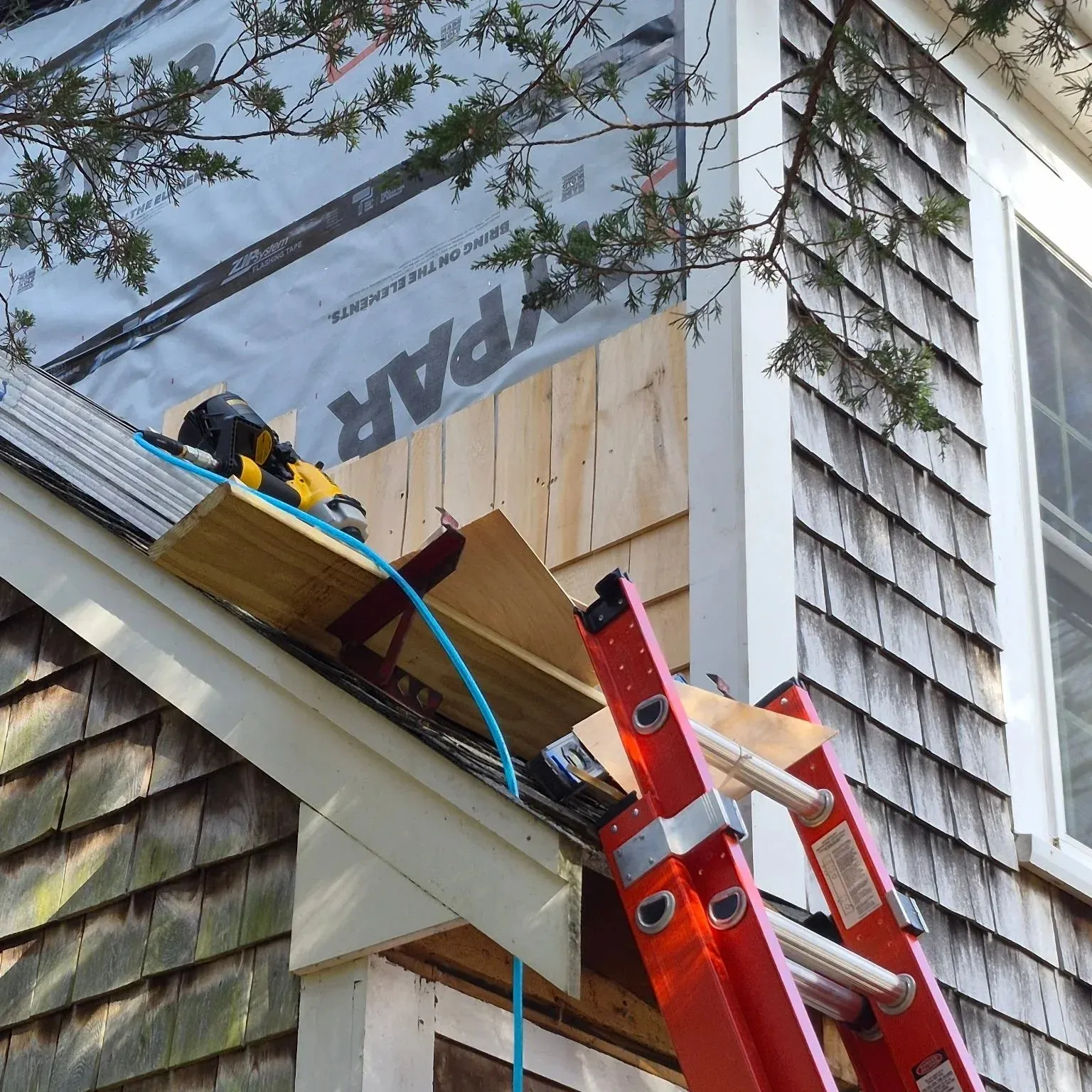 Exterior of a house under renovation with a leaning red ladder, construction tools, wooden panels, and partially installed siding.
