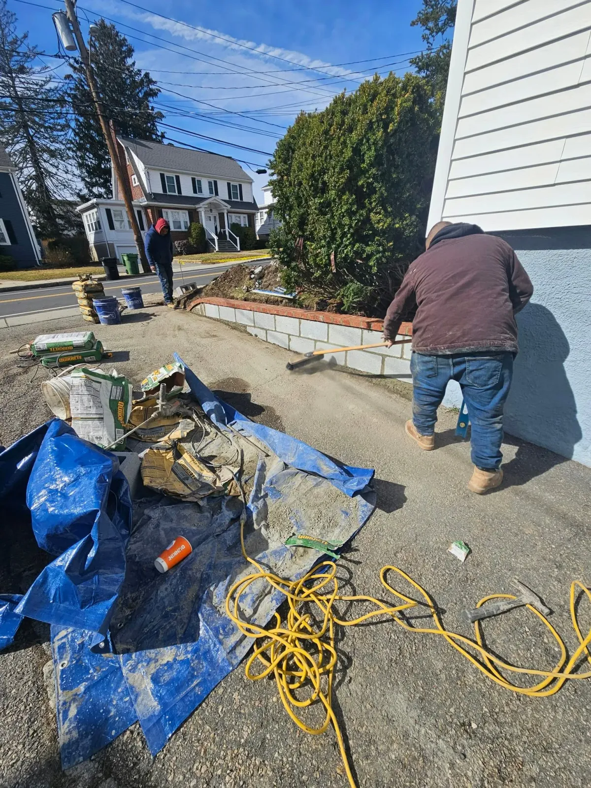 Worker laying pavers along a sidewalk beside a house, with tools, tarps, and extension cords nearby.