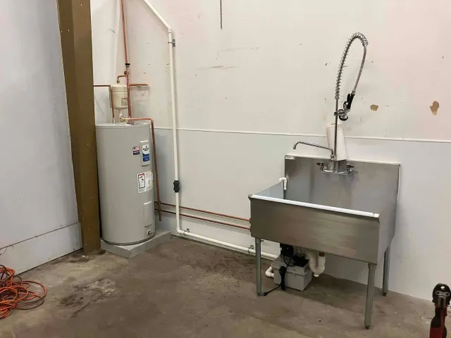 Utility room with a stainless-steel sink, water heater, and exposed pipes against a plain wall.