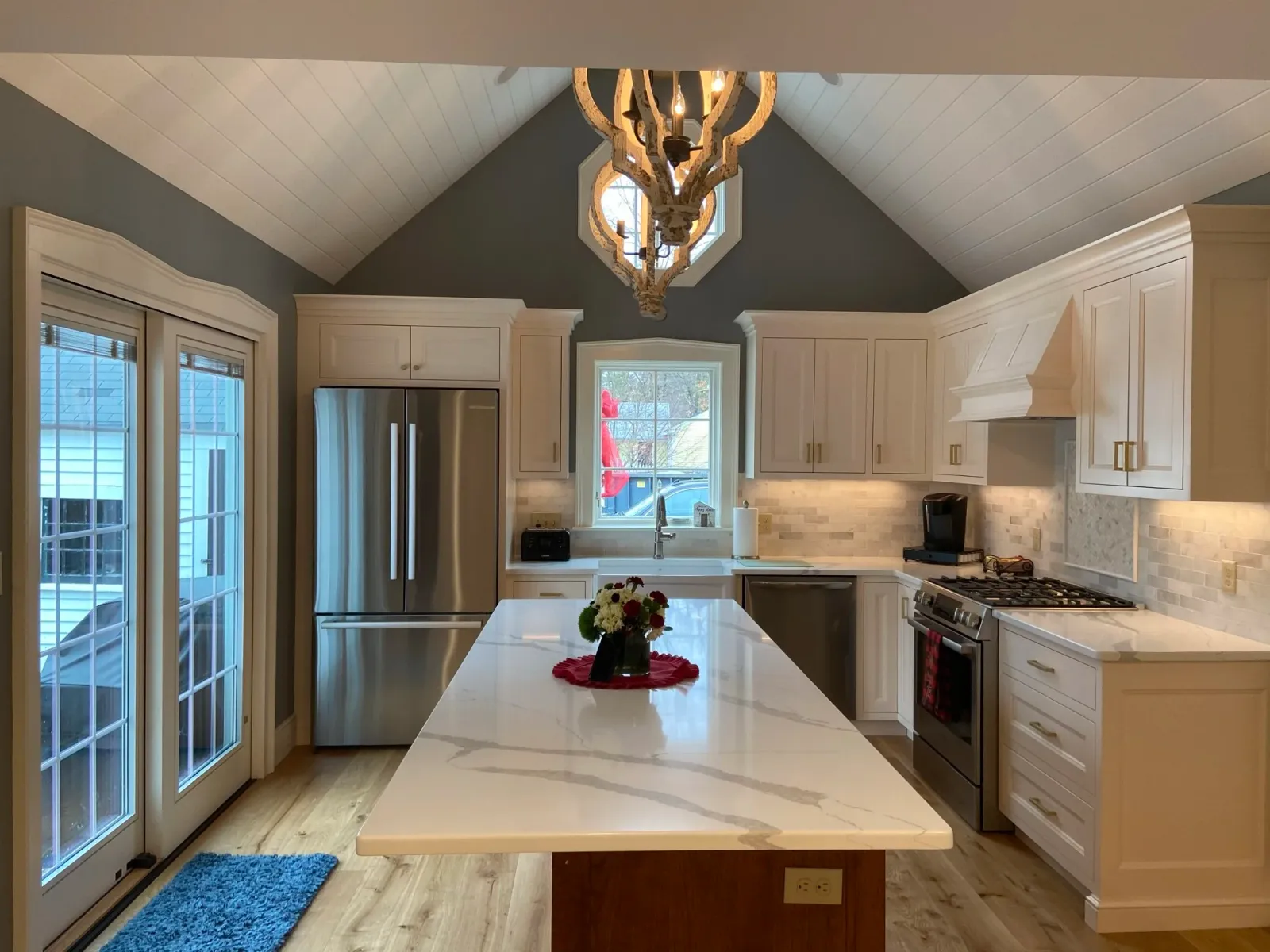 Modern kitchen with white cabinets, stainless steel refrigerator, gas stove, and a marble island counter, with double glass doors leading outside and a window above the sink. There is a chandelier hanging from the vaulted ceiling.