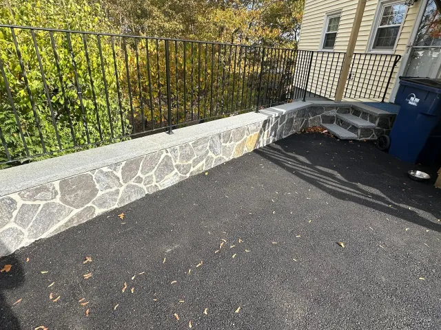 Stone retaining wall beside blacktop driveway with black railing, steps, and blue trash bin at right