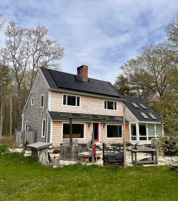 Two-story house with wooden siding and solar panels on the roof, surrounded by grass and trees. The house has a chimney, several large windows, and a patio area with outdoor furniture.