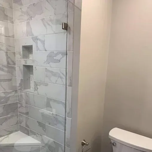 A bathroom with a glass-enclosed shower featuring large white and gray marble tiles, built-in shelves inside the shower, a partial view of a toilet, and a wall-mounted toilet paper holder.