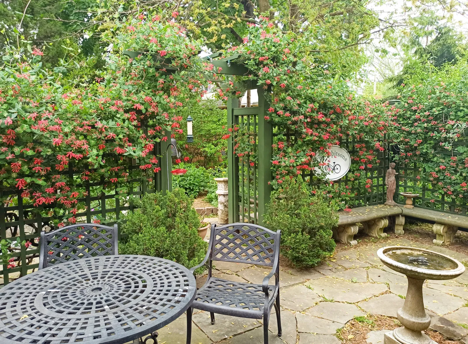 Garden patio with wrought iron furniture, trellis with climbing plants, and bird feeder.
