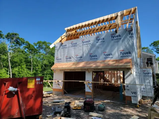 Construction of a two-story building with exposed wooden framing and Tyvek wrap. A red dumpster sits in front.