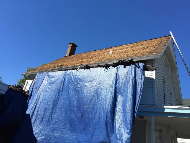 A house with a damaged roof partially covered by a large blue tarp against a clear blue sky.