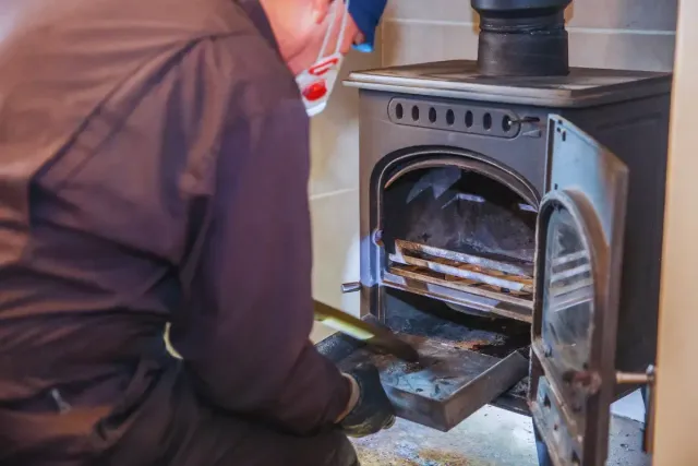 Person in mask cleaning a black wood-burning stove, removing ash tray. Stove door open.