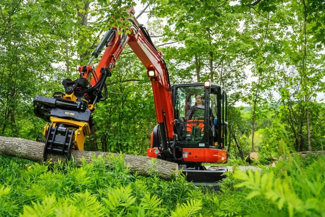 A person operating an orange excavator cutting down a fallen tree in a green forest.