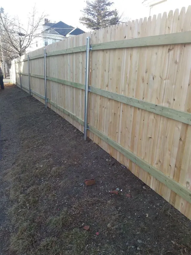 Wooden fence along a gravel path with silver poles for support. A few buildings are in the background.