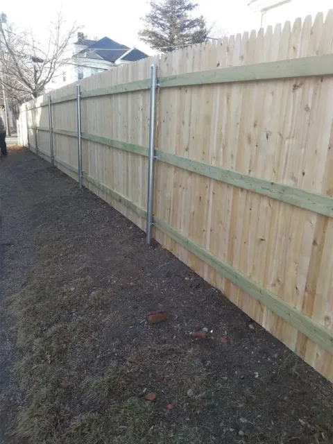 Wooden fence along a gravel path with silver poles for support. A few buildings are in the background.