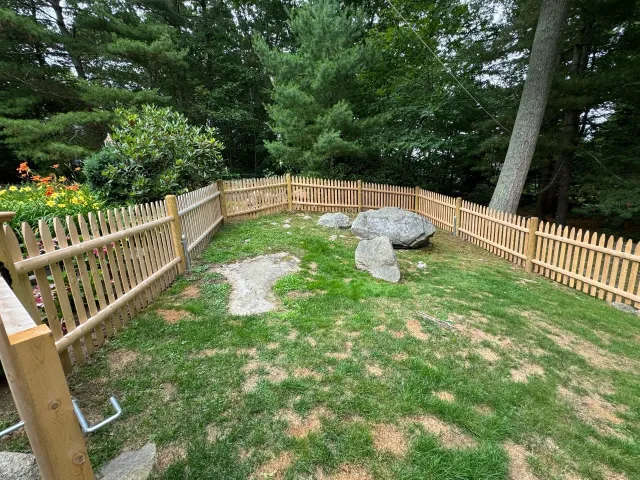 A wooden picket fence surrounds a grassy area with trees in the background.