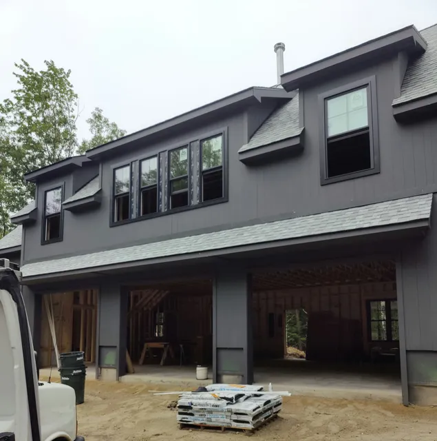 A gray two-story house under construction with several open garage doors. Construction materials are stacked in the foreground. Trees are visible in the background.