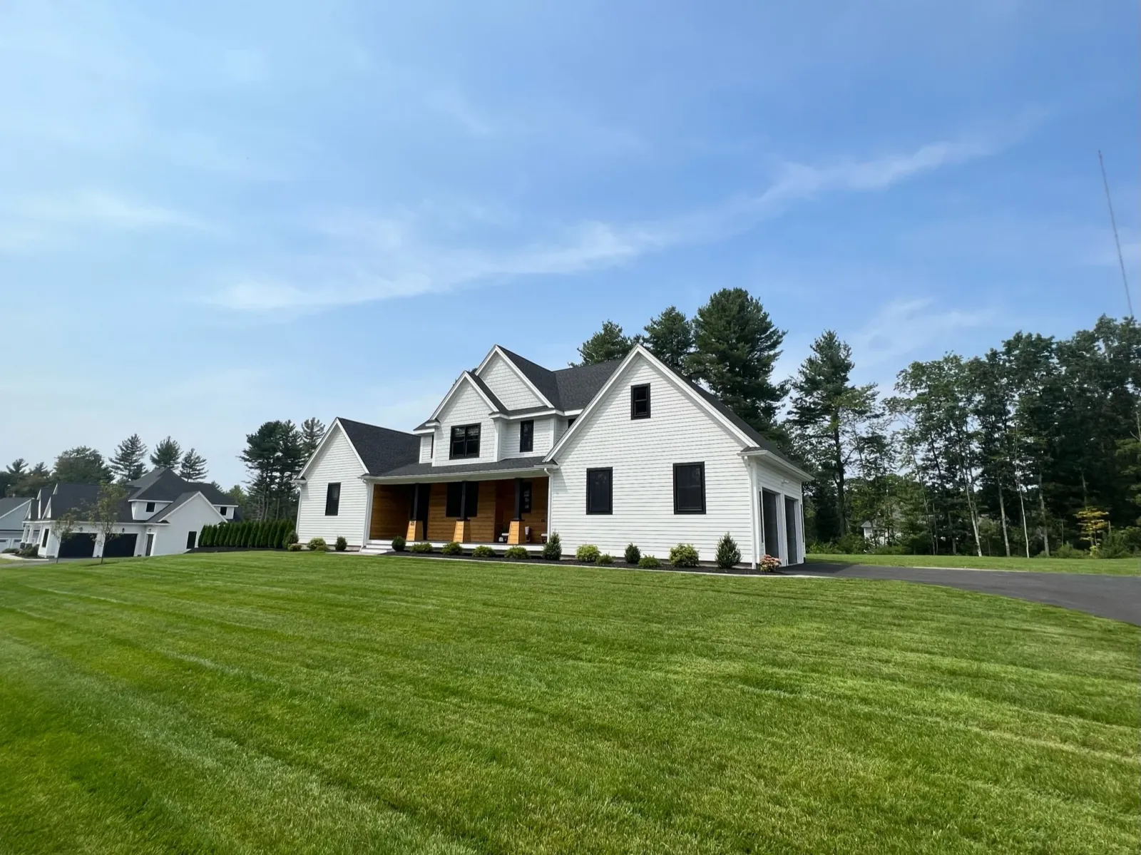 A white, two-story house with black roof and black window frames, featuring a large front porch with wooden accents, surrounded by a well-maintained green lawn and trees in the background under a blue sky.