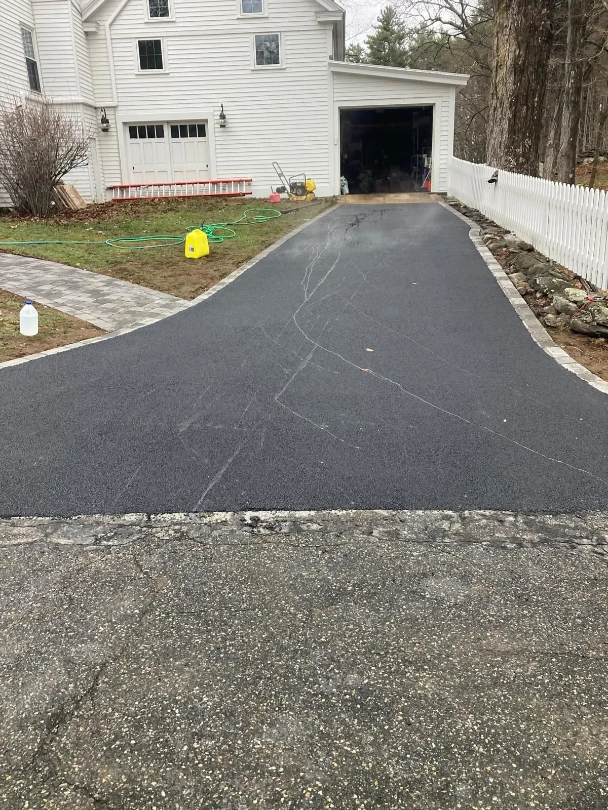 Freshly paved black driveway leading to a garage beside a white house on an overcast day