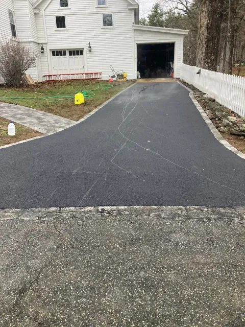 Freshly paved black driveway leading to a garage beside a white house on an overcast day