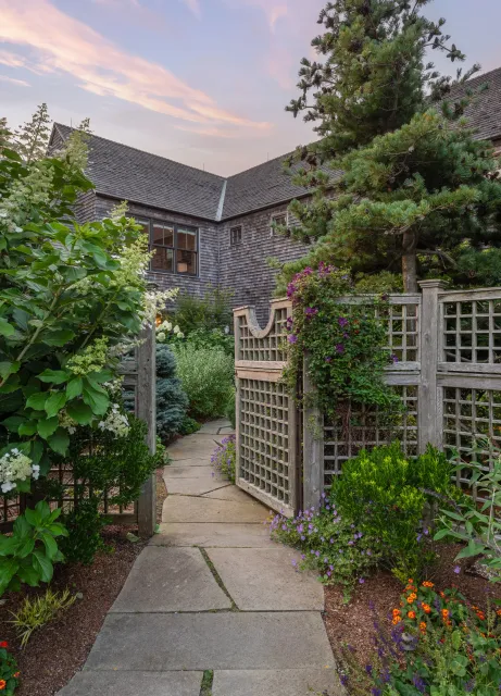 Pathway leading to a house backyard with wooden gate and garden, surrounded by lush green plants and colorful flowers, against a backdrop of a house with wooden shingles and a sky at sunset.