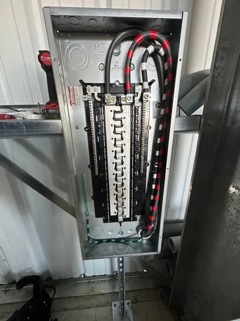 Electrical panel with black wires, installed on a metal bracket against a white wall.