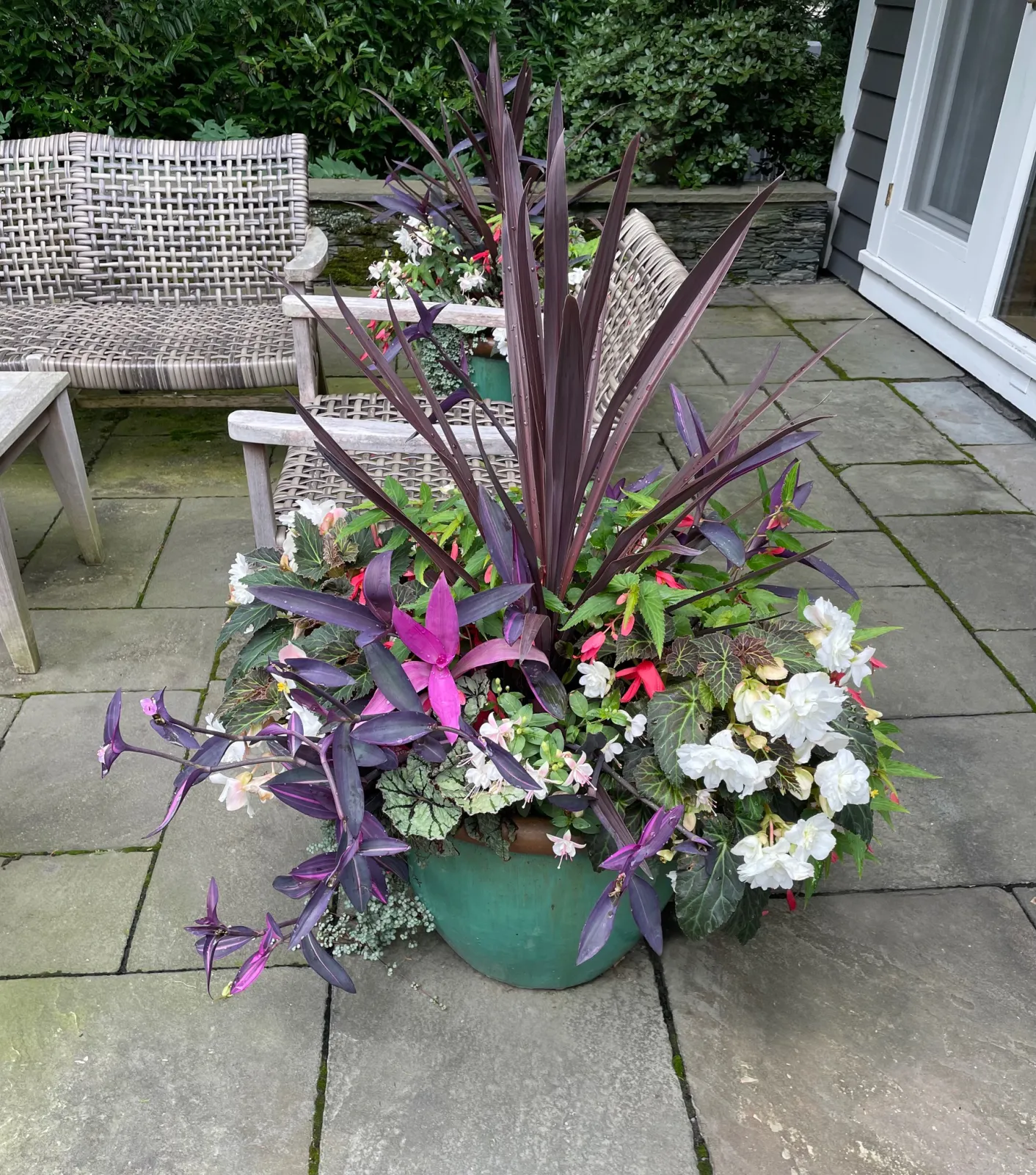 A green flowerpot filled with purple, white, pink, and green flowering plants is placed on a stone patio outside a house, with a gray and black siding wall and a white door visible in the background.