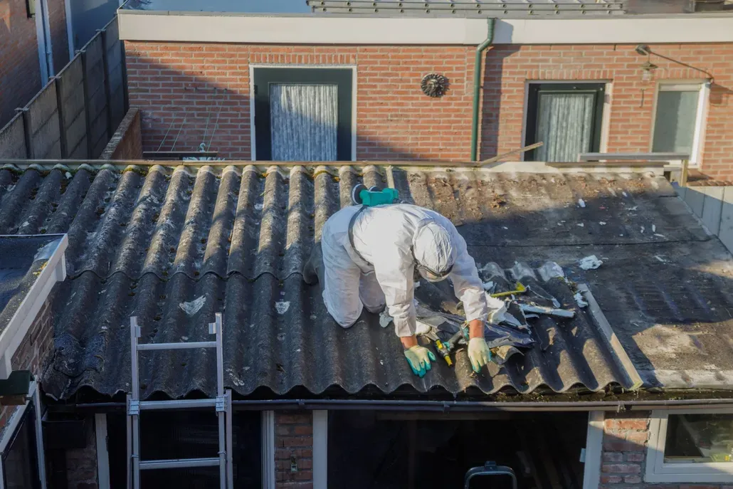 Worker repairing a corrugated roof on a brick building, with tools and ladder nearby.