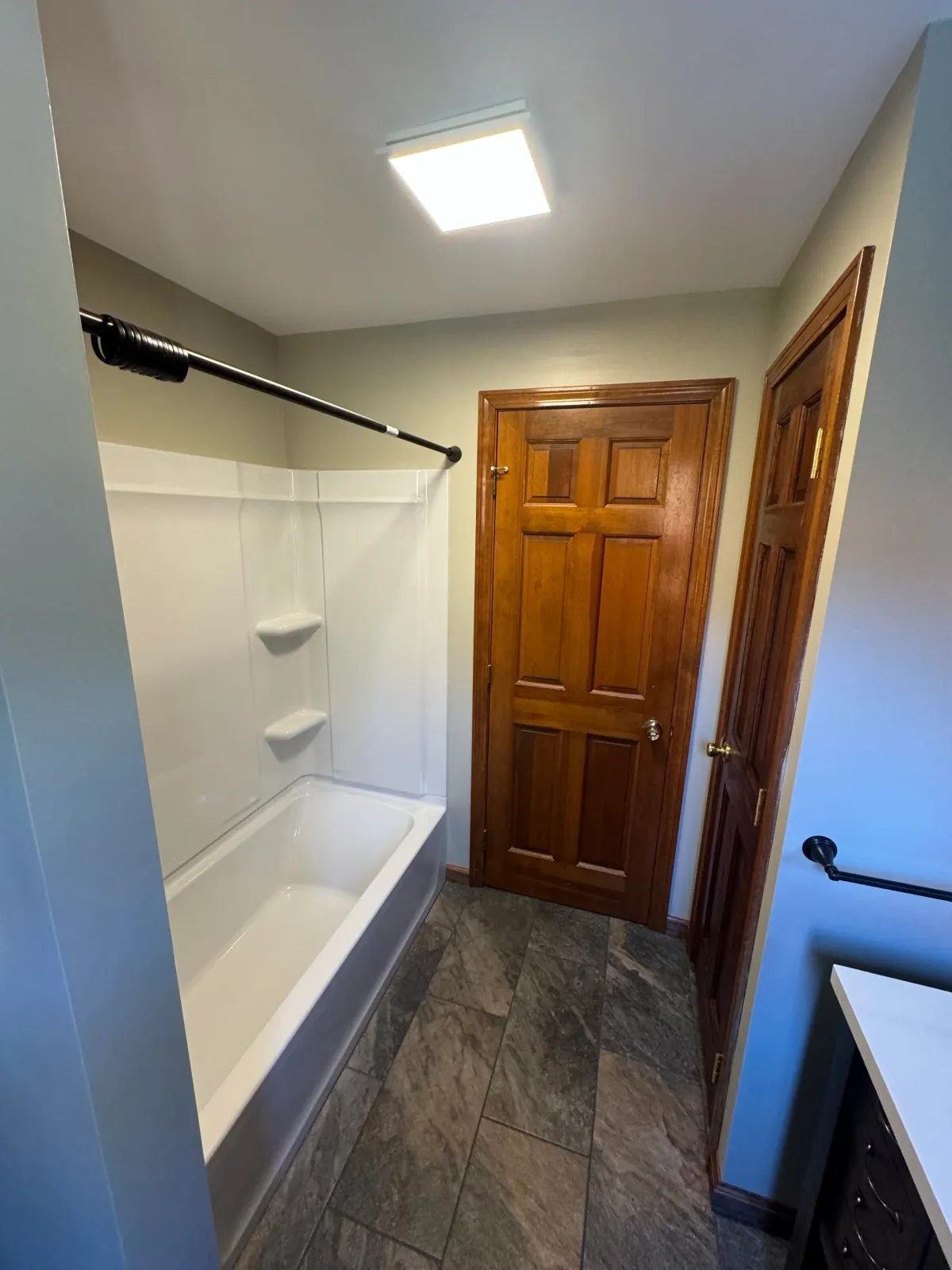 A small bathroom with a white bathtub, a shower curtain rod, a wooden door, and a partial view of a vanity with a black handle. The bathroom has a ceiling light and tiled floor.