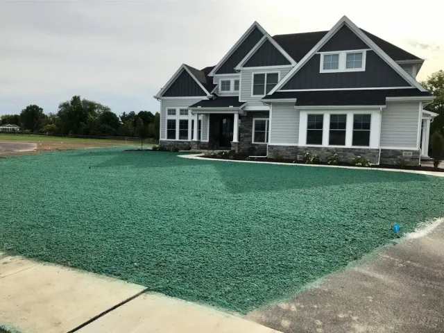 Newly constructed two-story house with dark and white siding, multiple gables, and large windows, siting behind a green newly hydroseeded yard and concrete sidewalk.