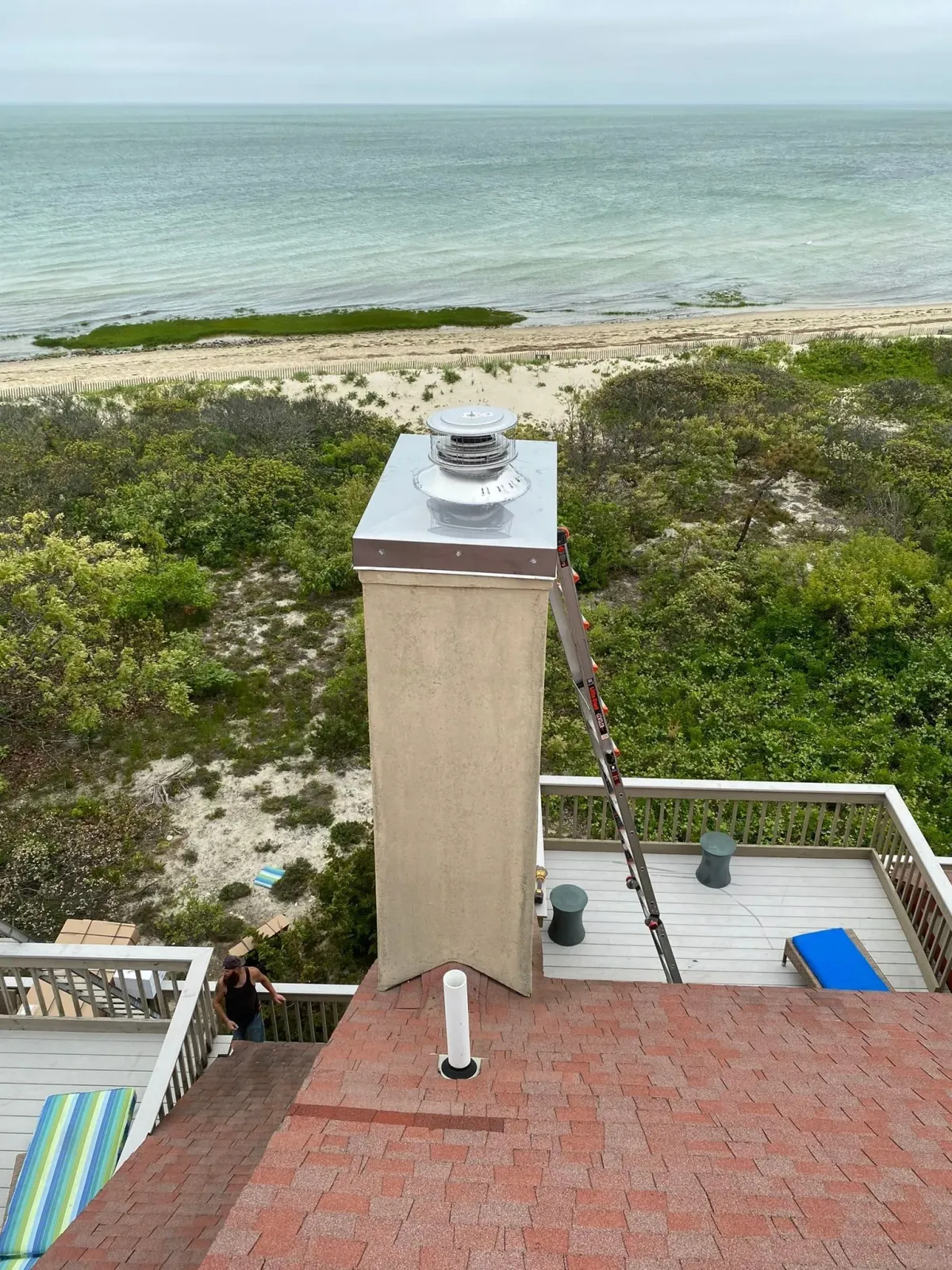 A chimney with a metal cap and a ladder leaning against it on a shingled roof overlooking a beach and the ocean.