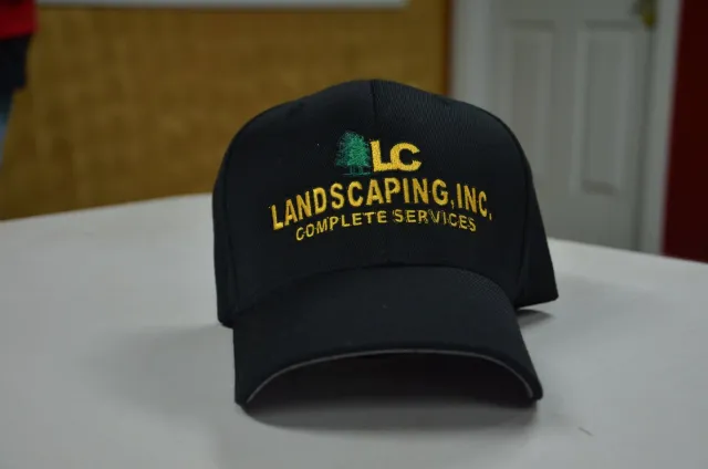 Black baseball cap with yellow embroidered text and a small green tree graphic, reading 'LC Landscaping Inc. Complete Services'.