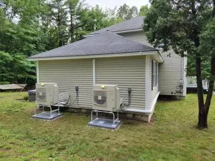 Two outdoor HVAC units beside a beige house with a gray roof and trees in the yard