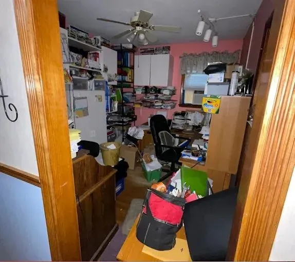 Messy, cluttered office/kitchen combo: piles of papers, appliances, and storage, with overhead lighting.