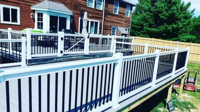 A newly installed backyard deck with black and white railings, attached to a red wooden house, with outdoor furniture and a green lawn surrounded by a wooden fence.